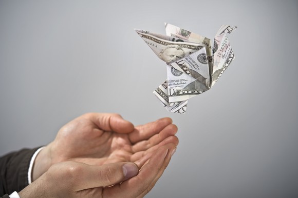 An origami bird made of U.S. paper currency flies away from a man's cupped hands.