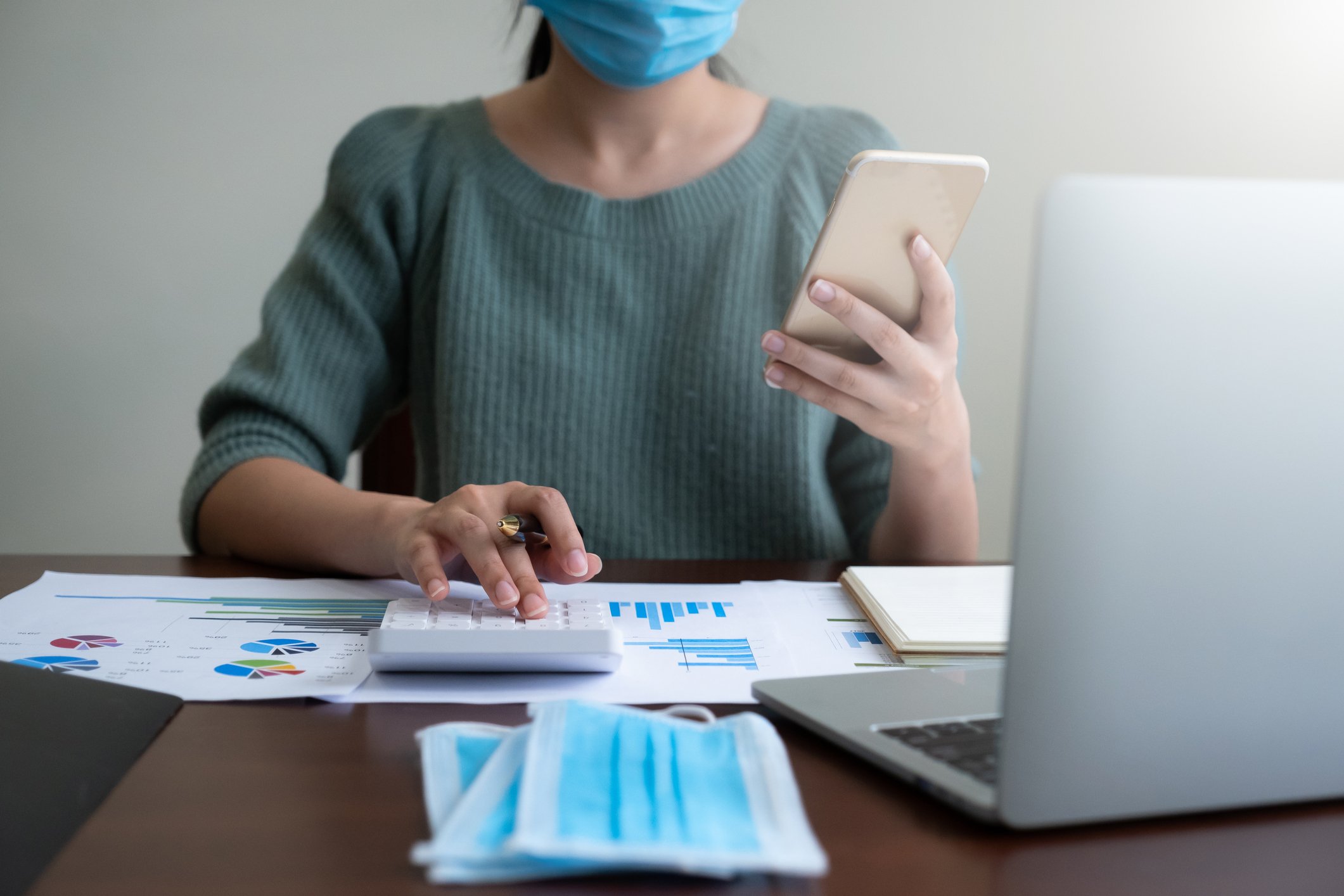 Woman wearing a mask while holding a phone in one hand and pressing buttons on a calculator with the other hand