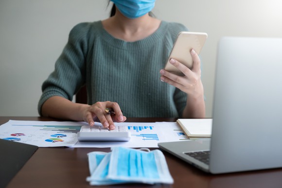 Woman wearing a mask while holding a phone in one hand and pressing buttons on a calculator with the other hand