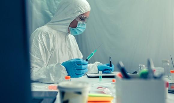 A scientist in protective gear takes notes while holding a full syringe.