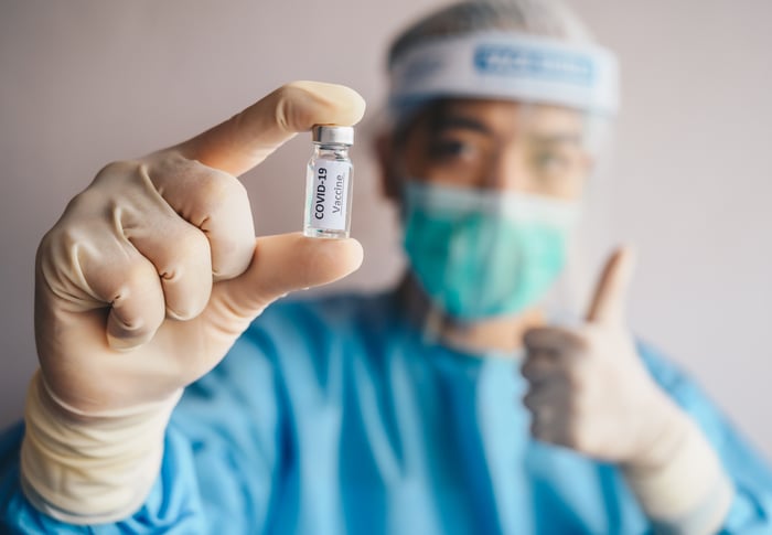 A scientist in protective equipment gives a thumbs-up while holding a vial labeled COVID-19 Vaccine