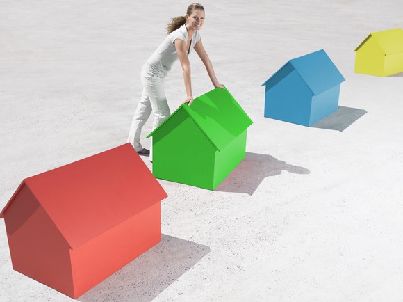A woman dressed in white leaning forward on a small green house, which is in a row of multicolored small houses. 