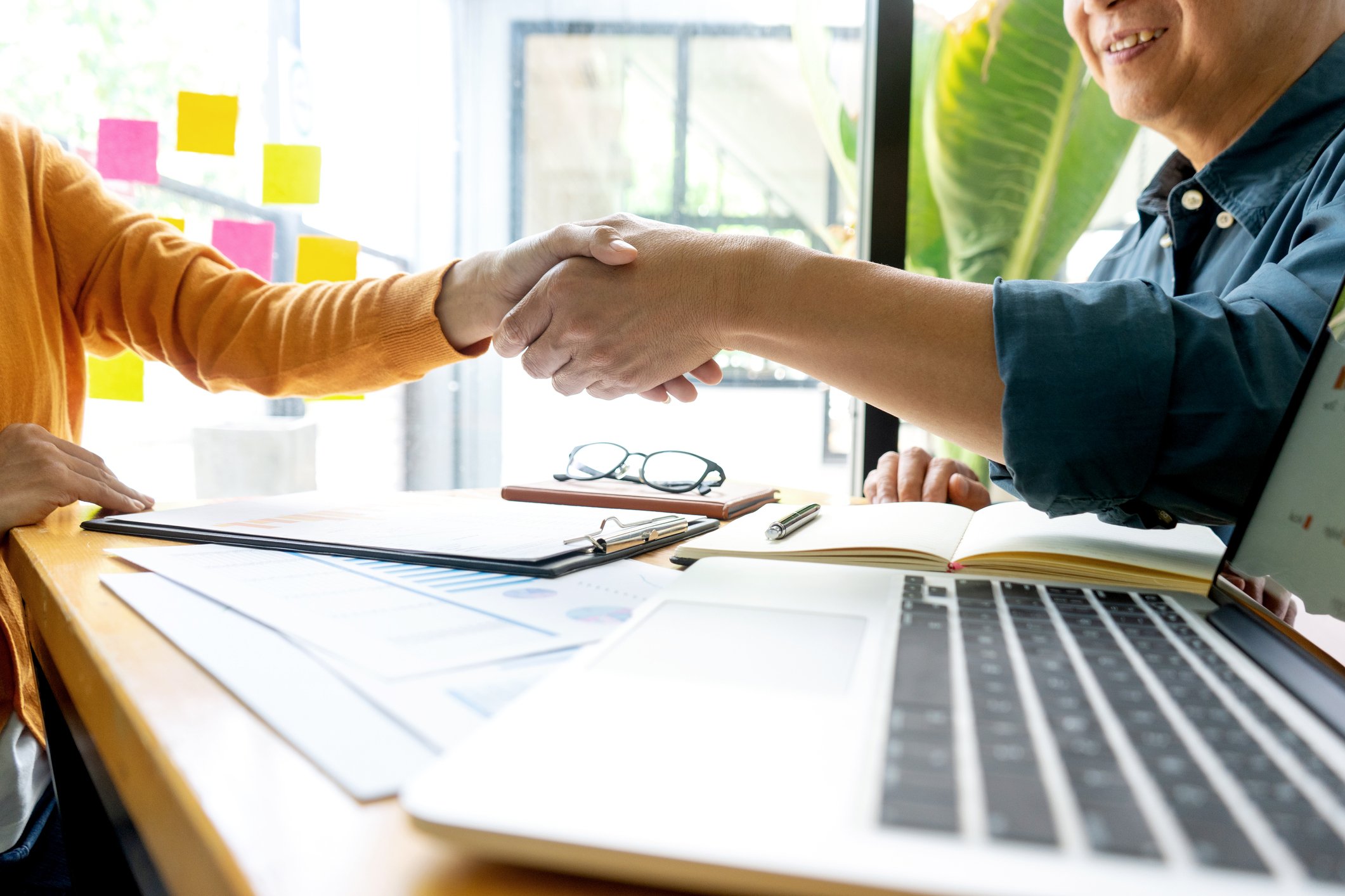 A smiling businessman shakes hands across a table with a woman in a yellow sweater, with an open laptop in the foreground