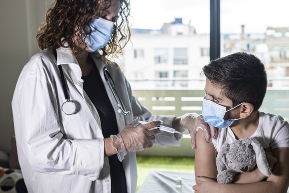 A healthcare worker administers a shot to a young boy. Both are wearing masks. The child is holding a toy bunny.