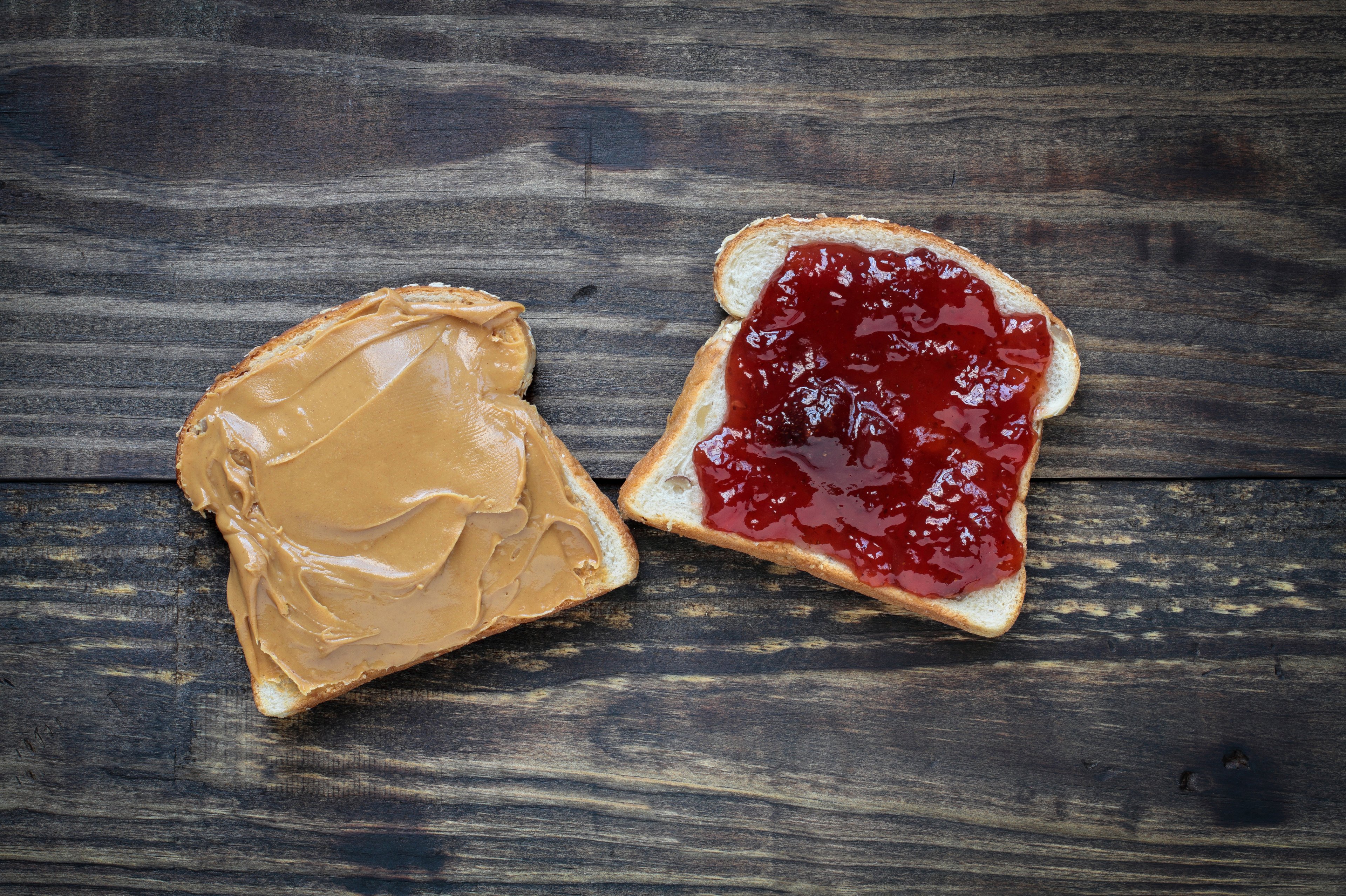 Two pieces of bread, one covered with peanut butter and the other with jelly, sit side by side on a wooden surface.
