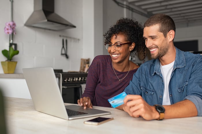 A young couple smile at the laptop as the man holds a credit card in their kitchen. 