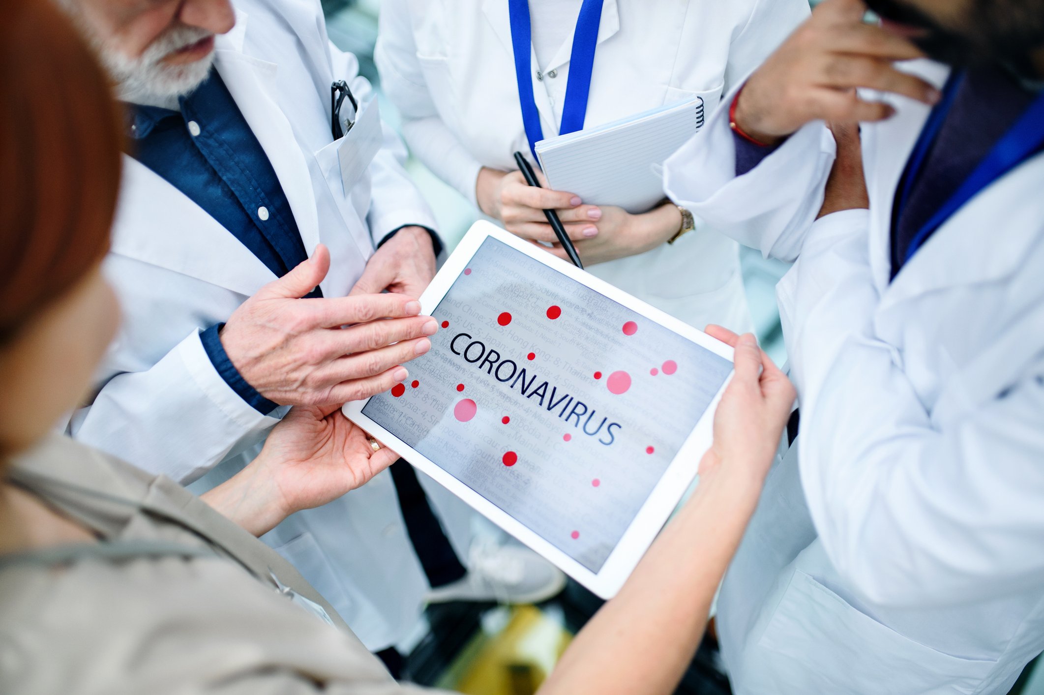 Four people wearing white coats standing in a circle around a touchscreen tablet displaying "coronavirus"