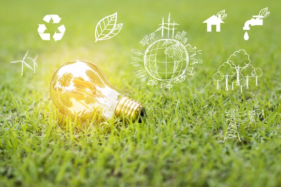 A glowing light bulb lies on the grass surrounded by renewable energy icons.