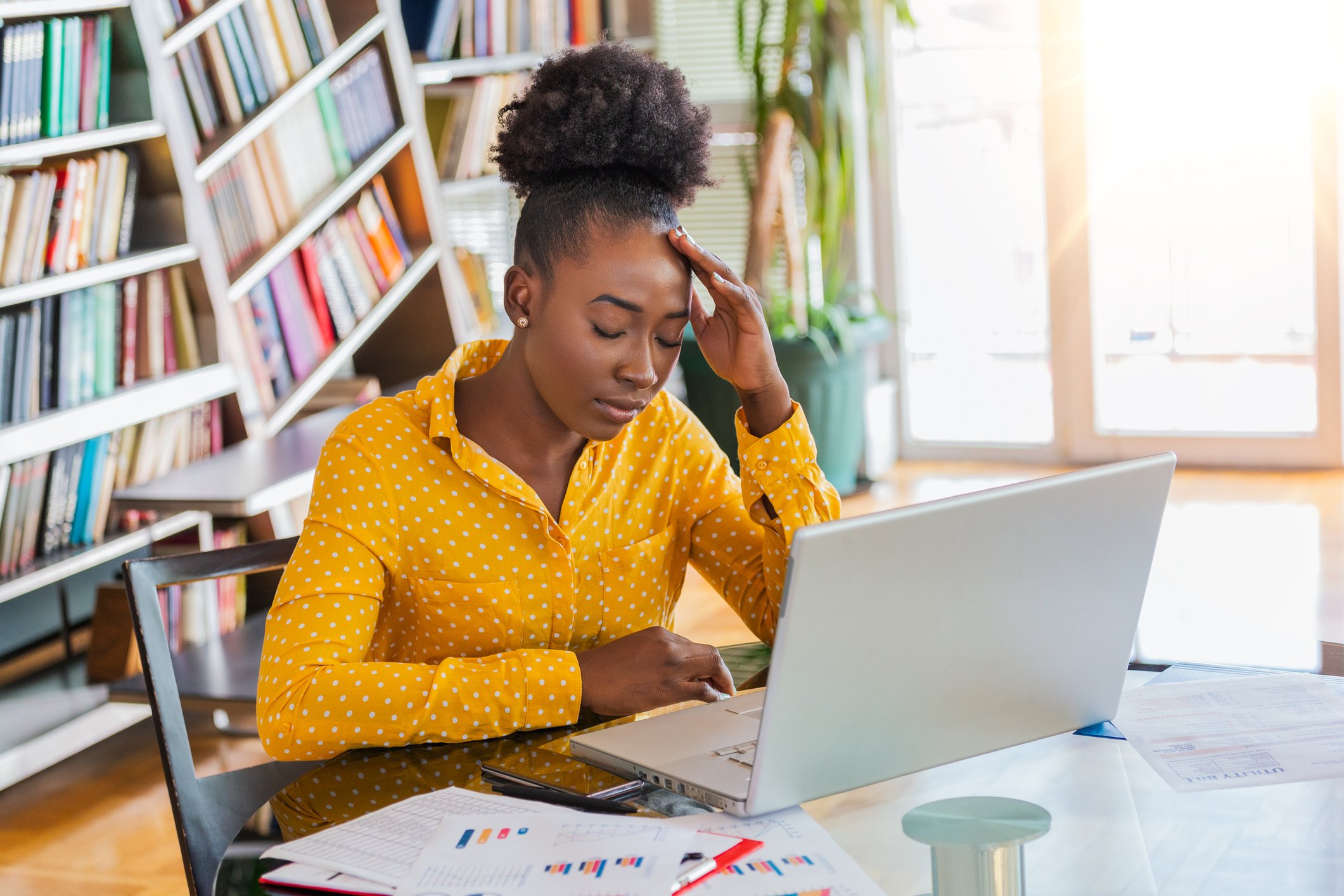 A young woman sits at a laptop with her eyes closed and her hand to her head.