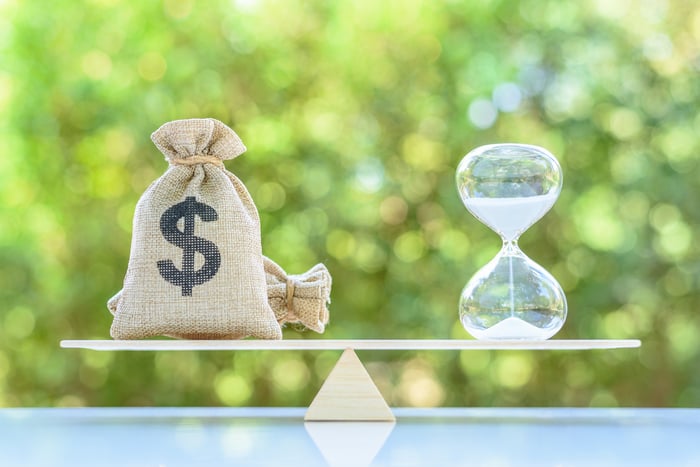 A bag with a dollar sign on it sits balanced on a scale with an hourglass.