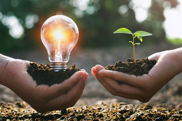 two hands holding dirt with light bulb and green plant sprout indicating clean energy