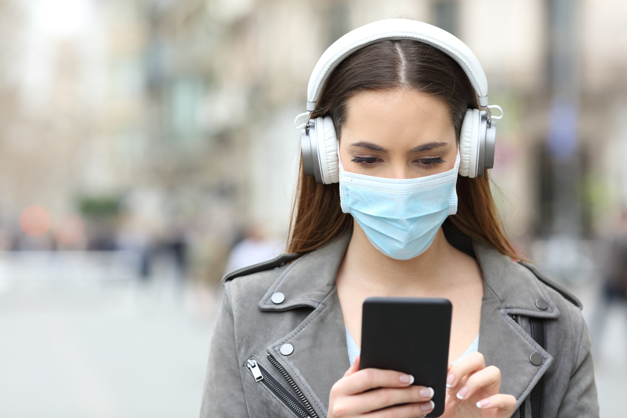 A young woman uses her smartphone, wearing a surgical mask and white headphones.