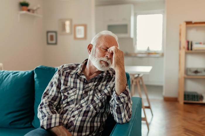 Older man sitting on the couch pinching the bridge of his nose with his fingers. 