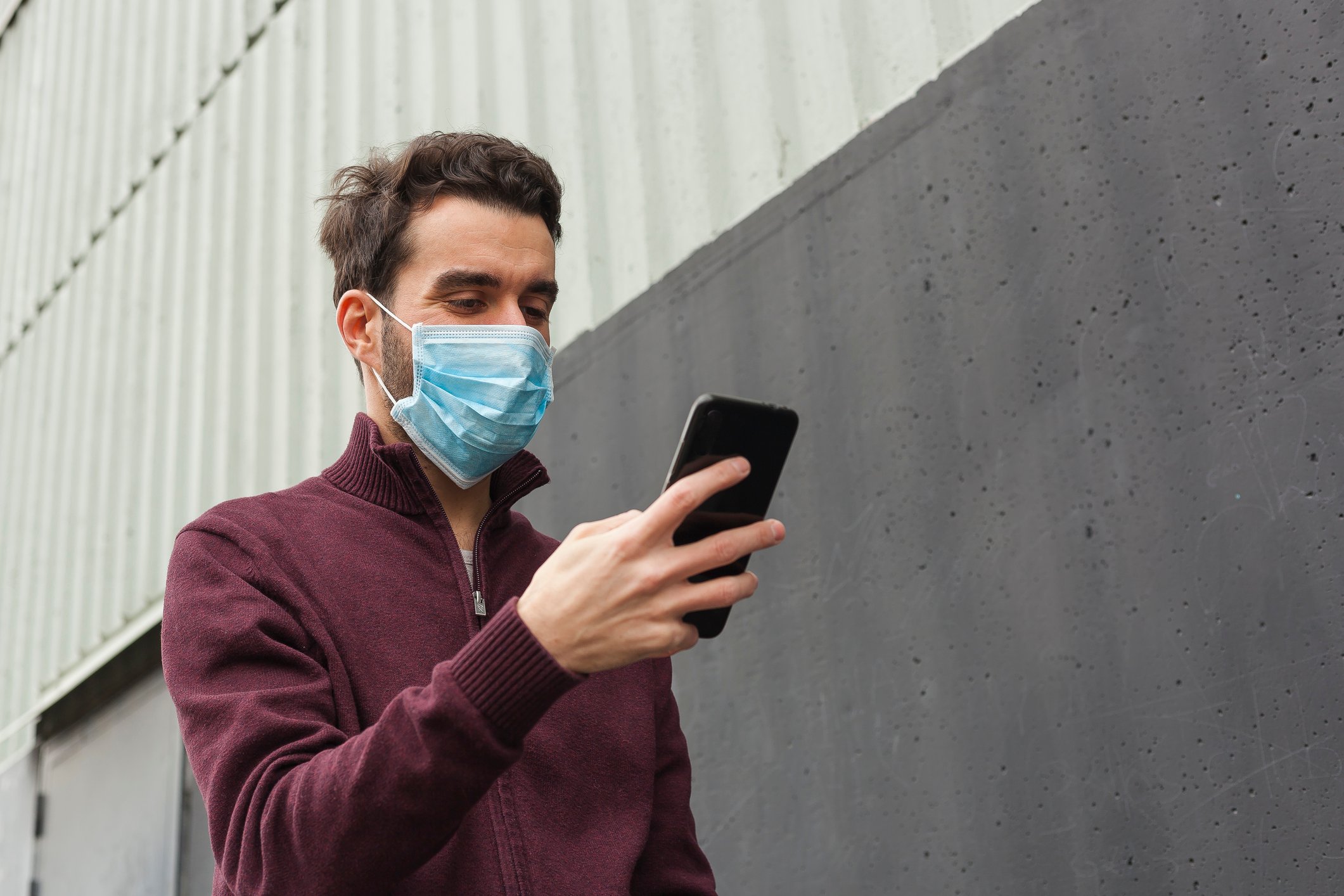 A masked man looks at his smartphone while walking beside a building.
