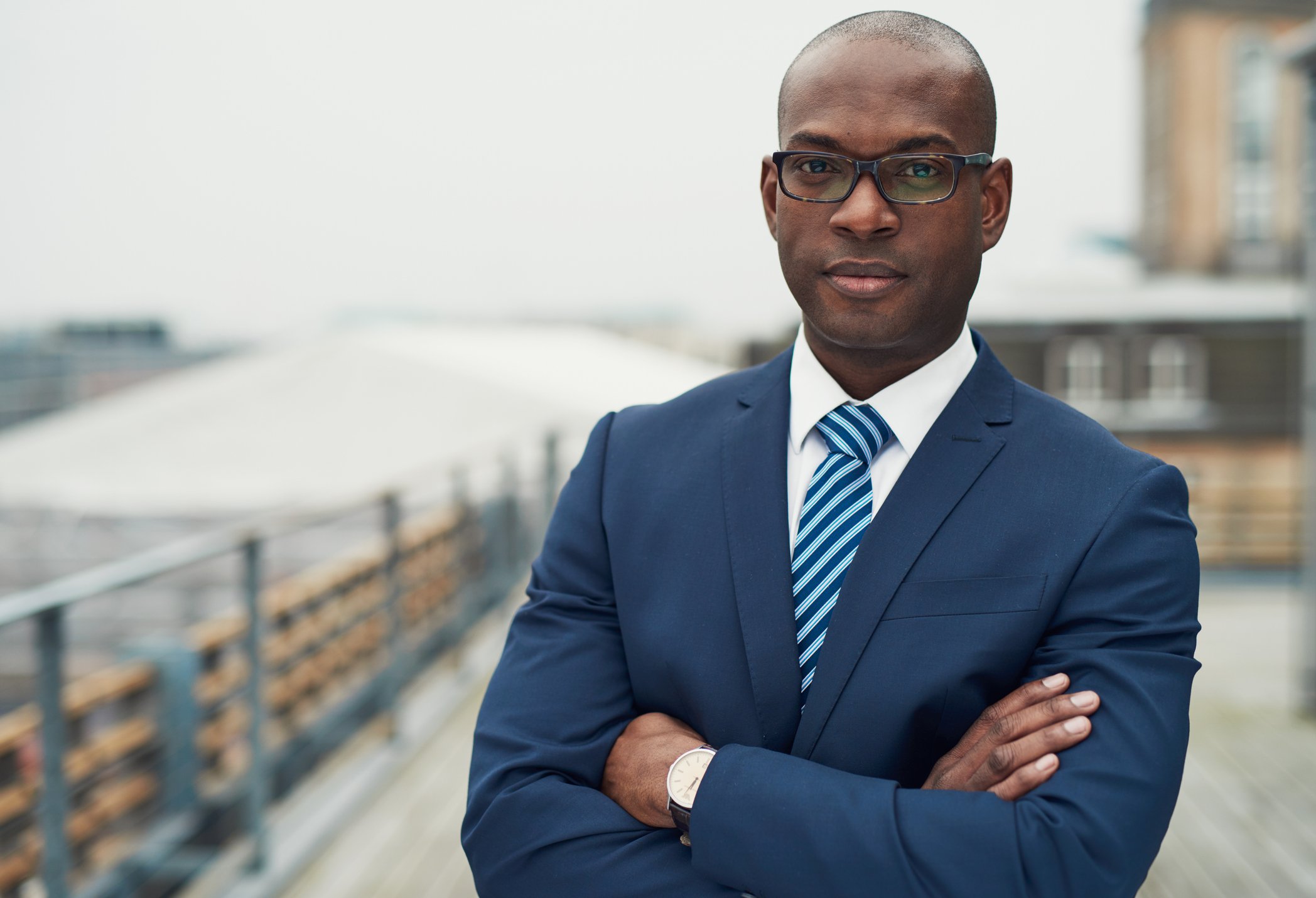 A man wearing a suit and tie stands with arms crossed on the roof of a building. 