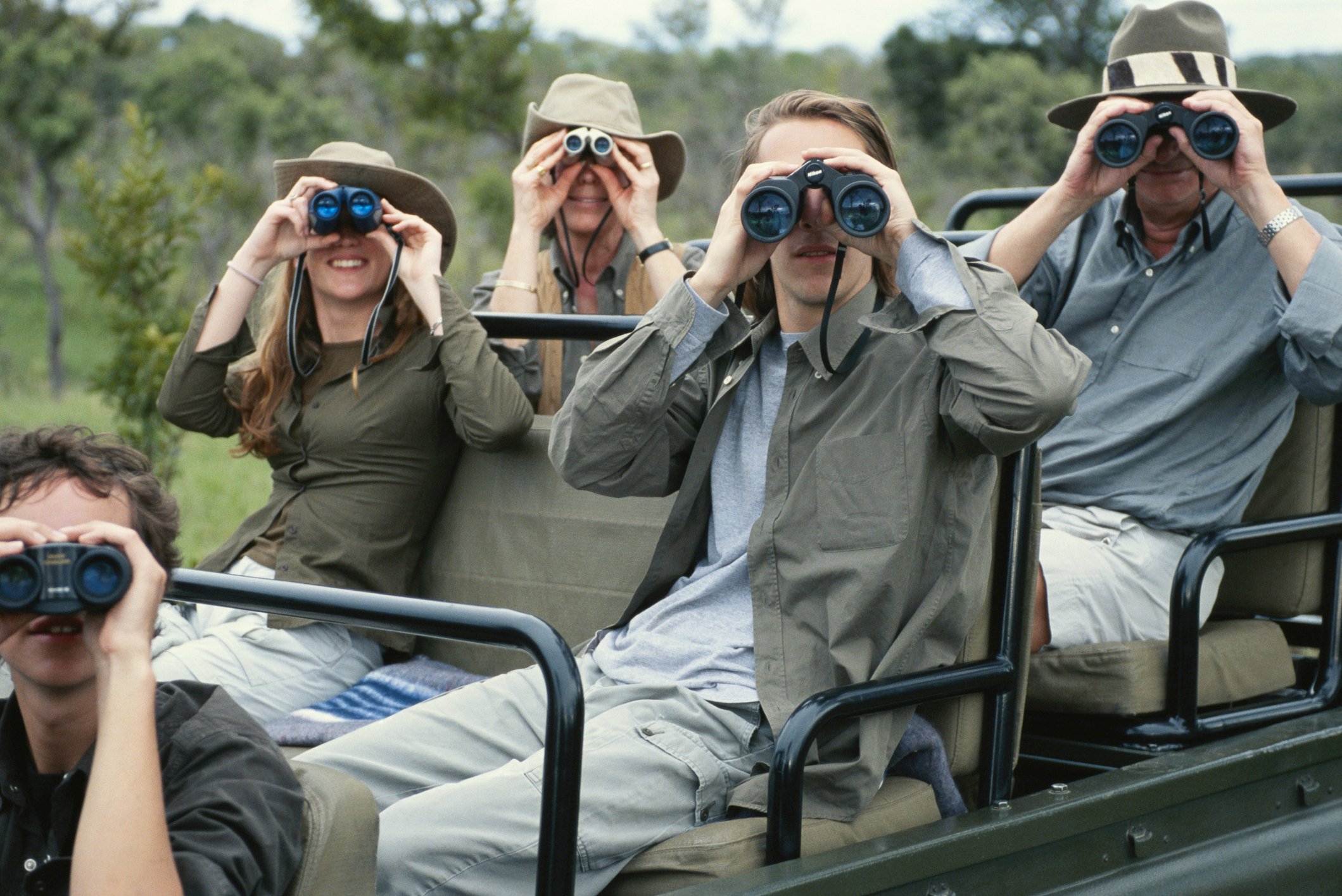 A traveling family sits on a Jeep and looks at camera with binoculars. 