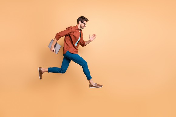 A young man carries a laptop and runs  in mid air against a beige background. 