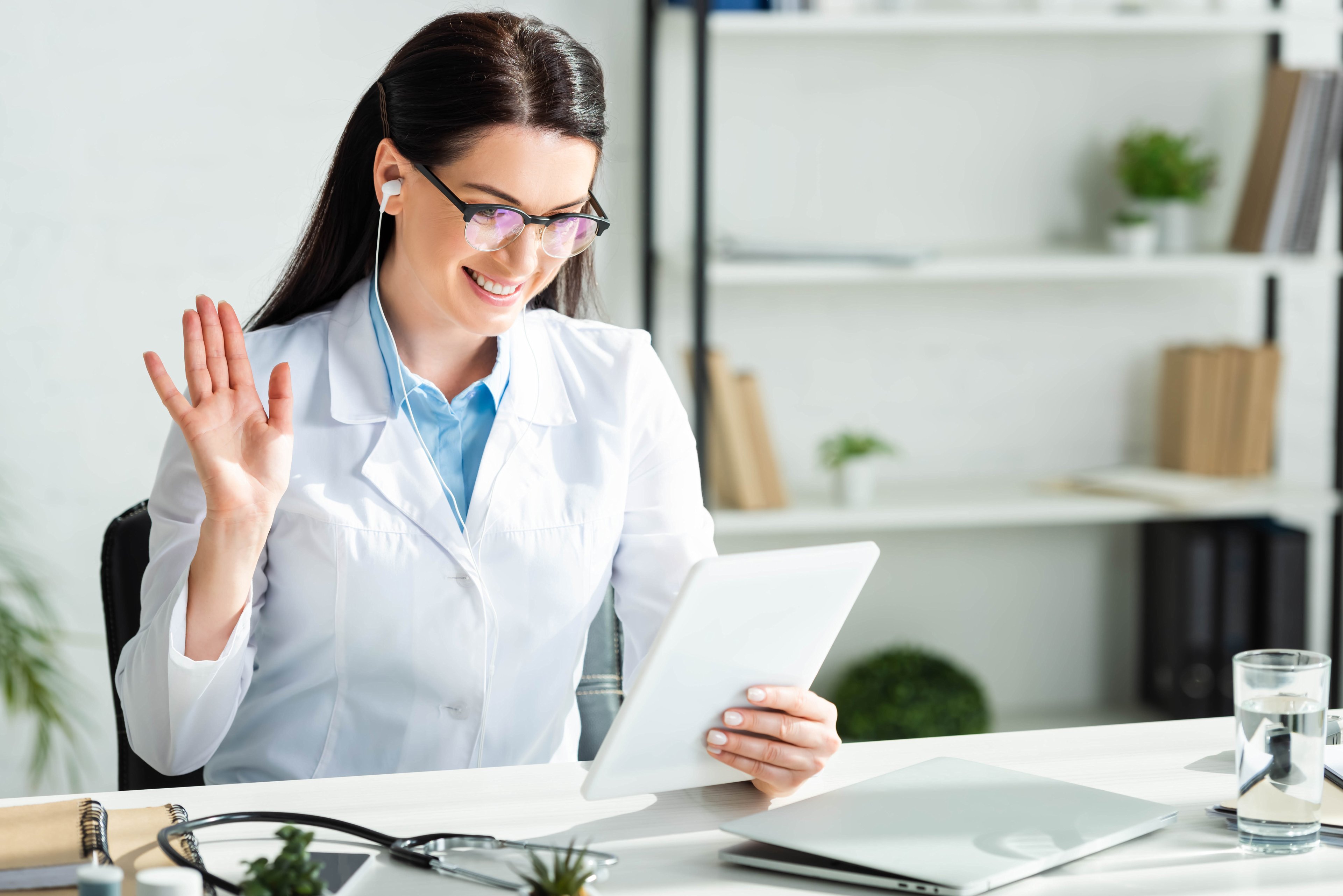 Doctor waving and having online consultation on a digital tablet in clinic office with laptop.