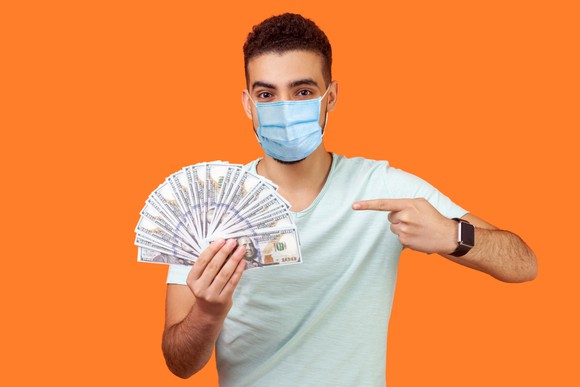 Man in mask holding hundred dollar bills and pointing to them with his other hand.