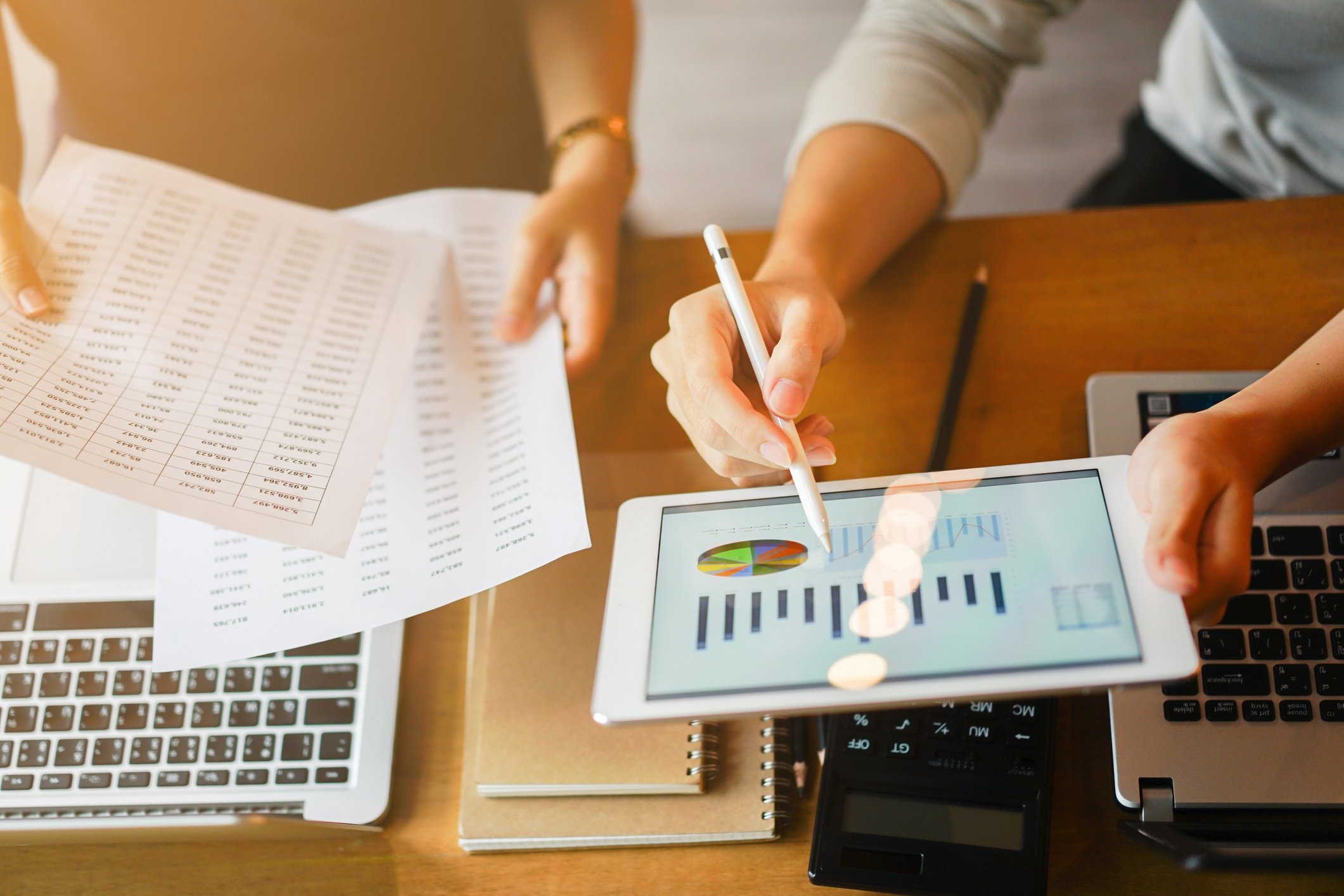 A person in an office holding a tablet displaying charts, next to another person holding papers with columns of figures