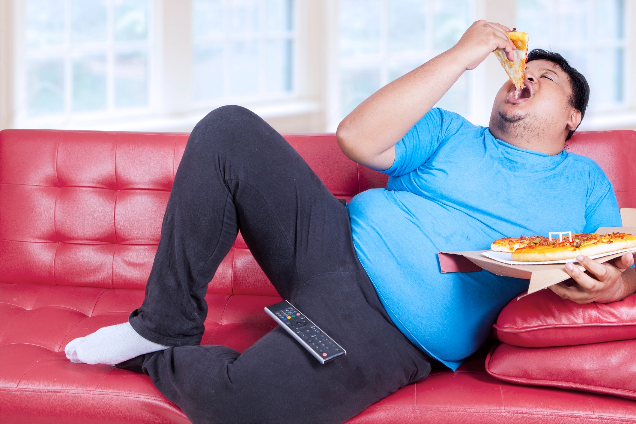 A man lounges on  the couch holding a full pizza and bites into a slice with the remote control on his thigh. 