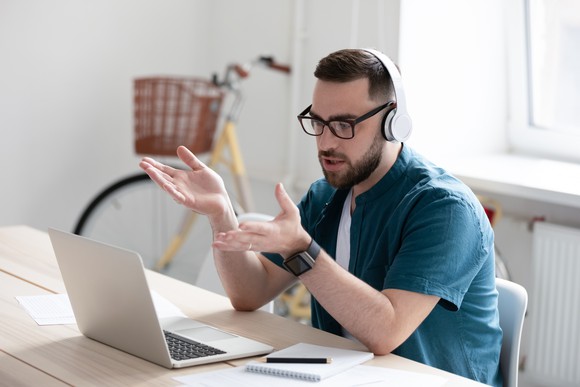 A man sits at a table with a laptop in front of him, wearing headphones and gesturing at the screen