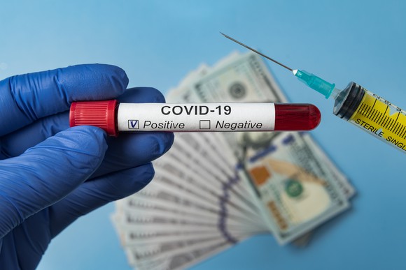 A gloved hand holding a vial labeled "COVID-19 positive" alongside a syringe and some American paper currency in the background.