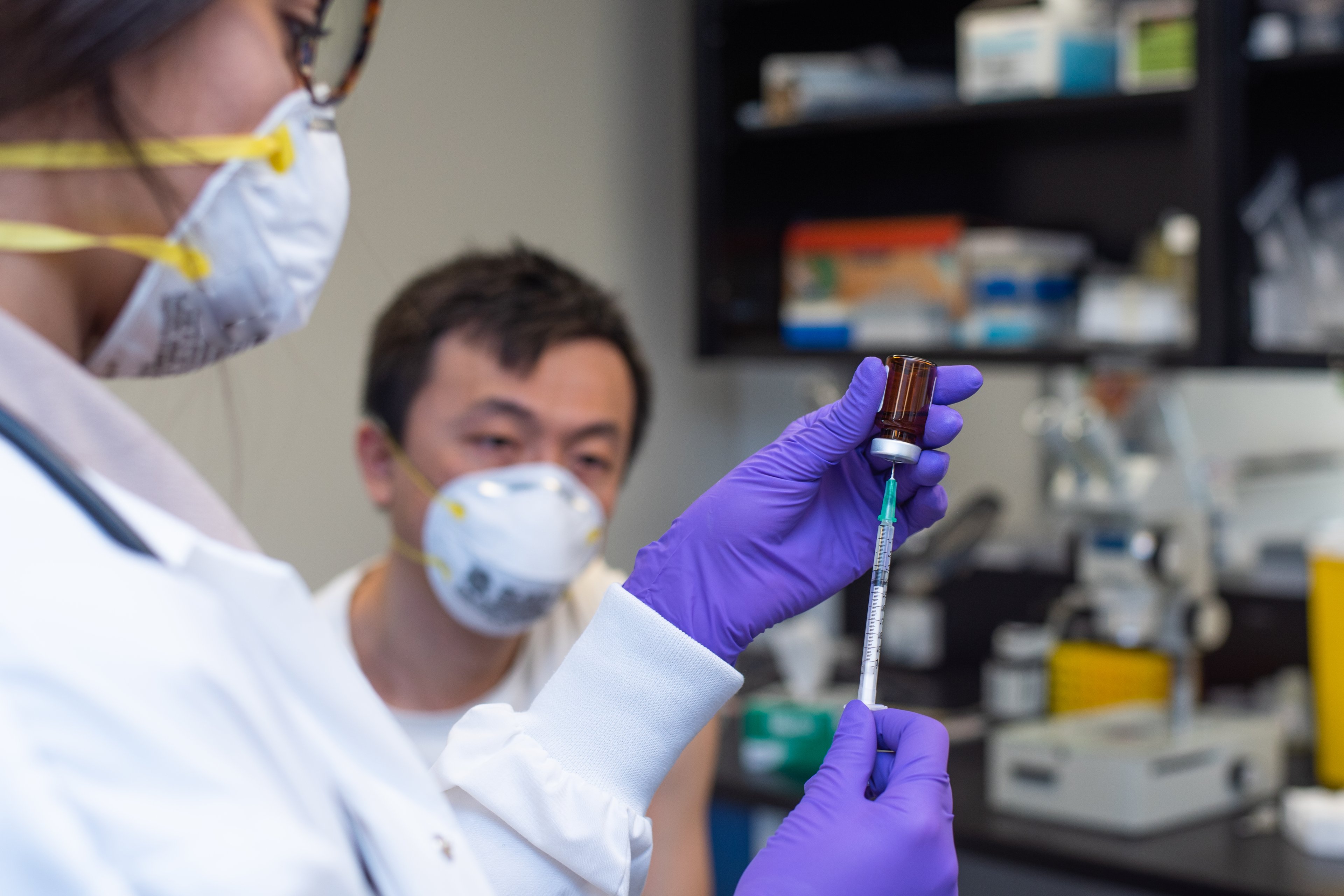 A masked medical professional draws medicine from a vial as a  masked patient sits nearby. 