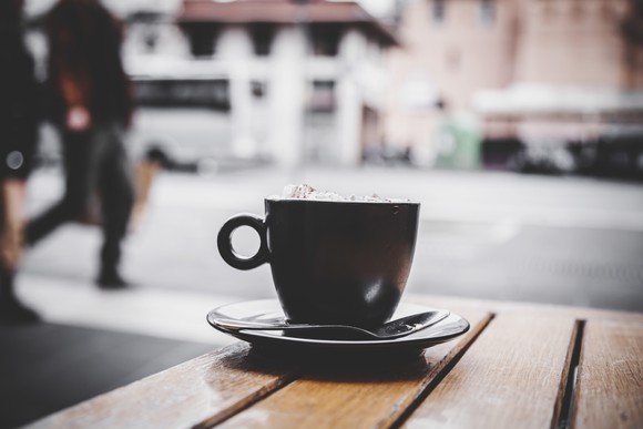 A cup of coffee sitting on a plate with a spoon on a wooden table outdoors.
