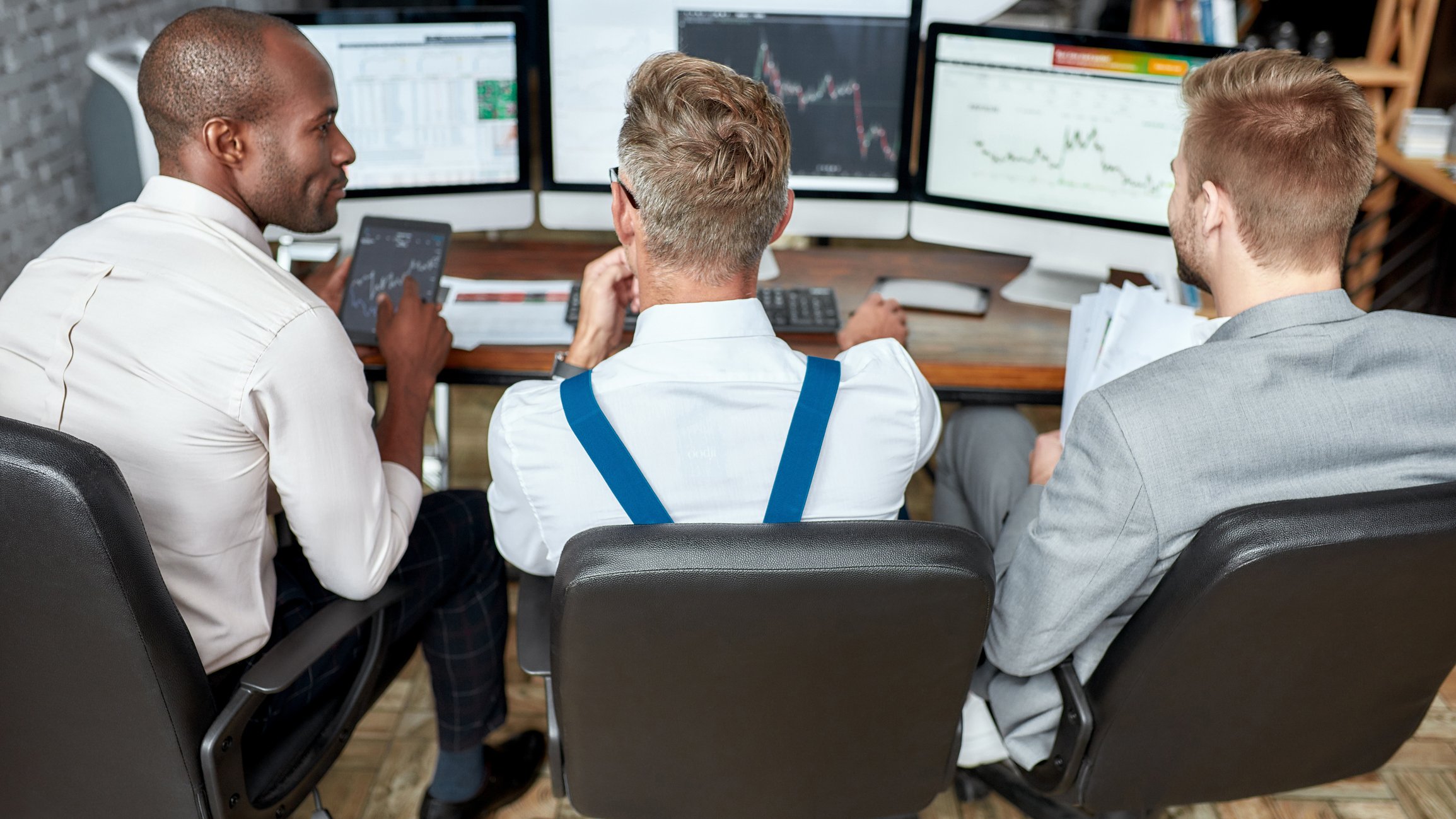 Three people gather around computers displaying stock charts.
