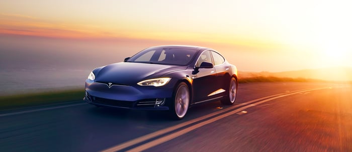 Blue Tesla Model S car on a road with sunset and water in background.