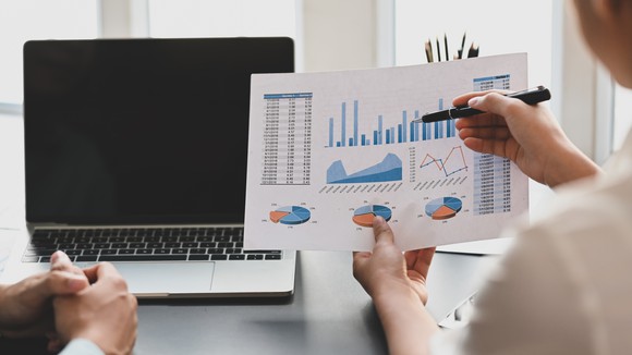 Woman explaining stock market data chart to man with a laptop at desk