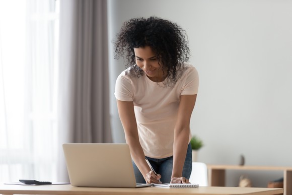 A woman stands, with notepad and pen, behind a laptop on a desk.