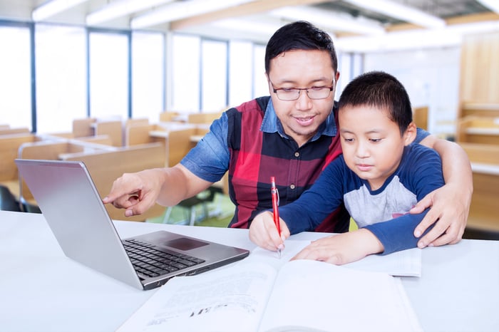 A man watching a little boy write and pointing to a laptop screen