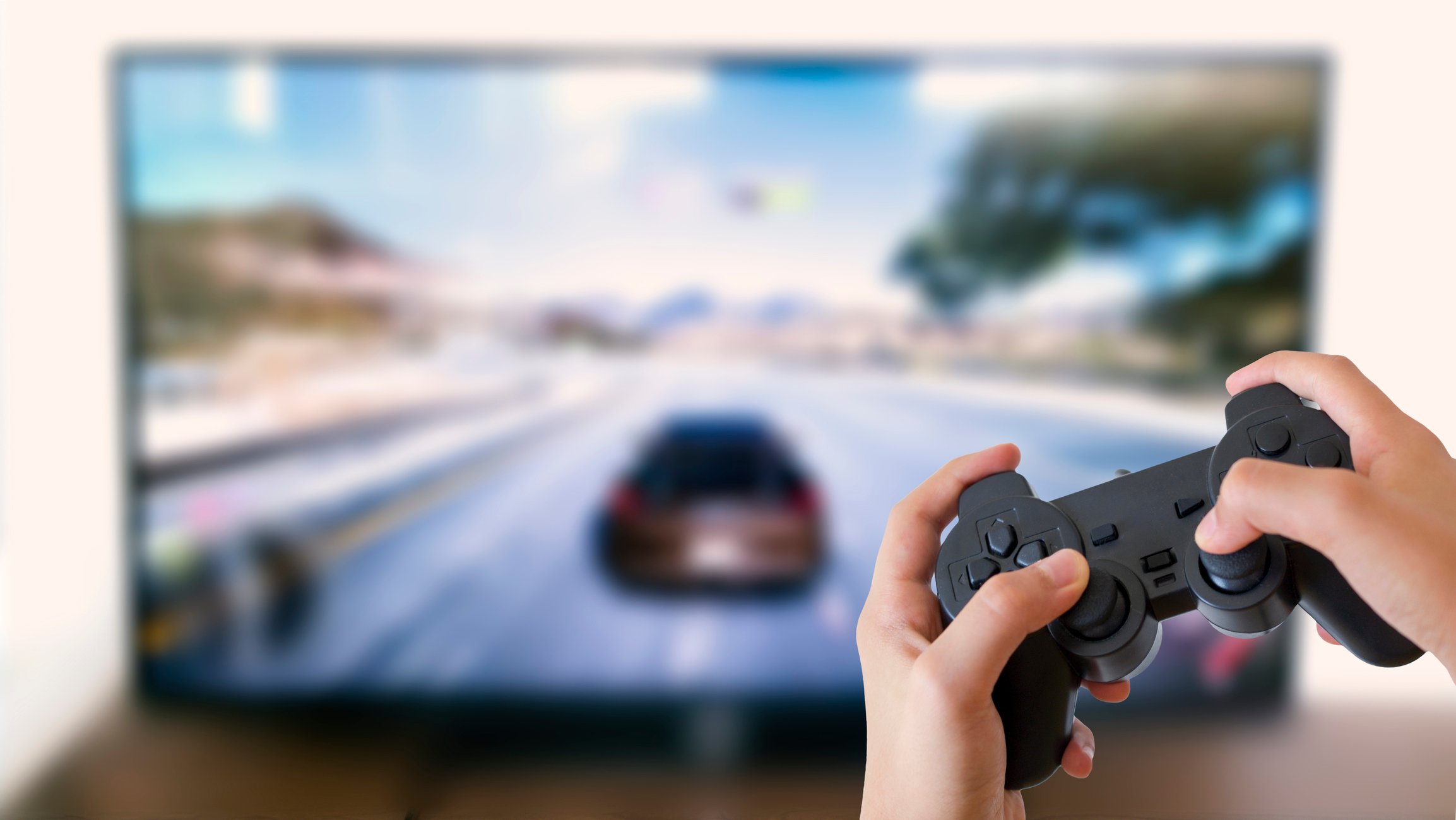 A hand holding a video game controller with a game displayed on a TV in the background.