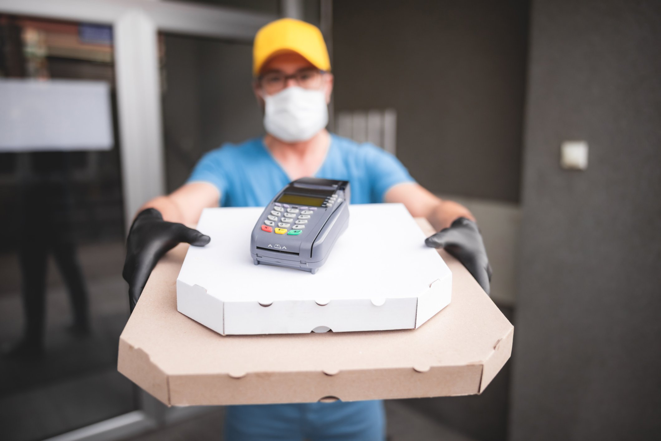 A person in a mask and gloves presenting a couple of boxes with a credit card scanner on top.