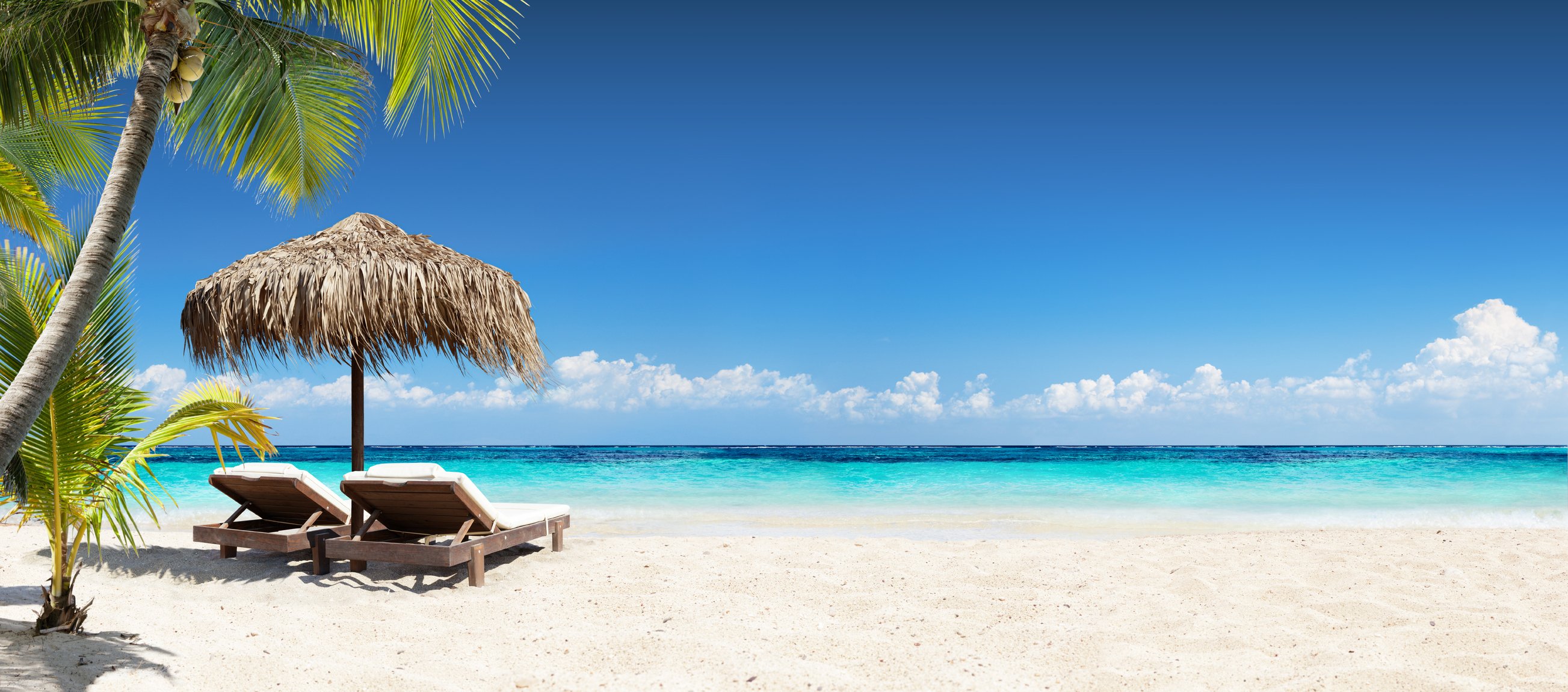 A beach with pale sand and two chaises set up under a thatched umbrella
