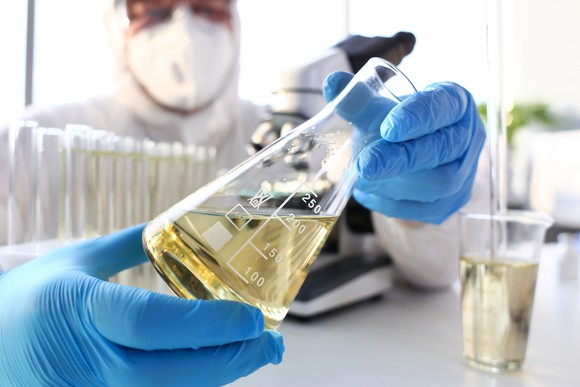 A masked scientist holding a bottle of yellow liquid in a laboratory. 