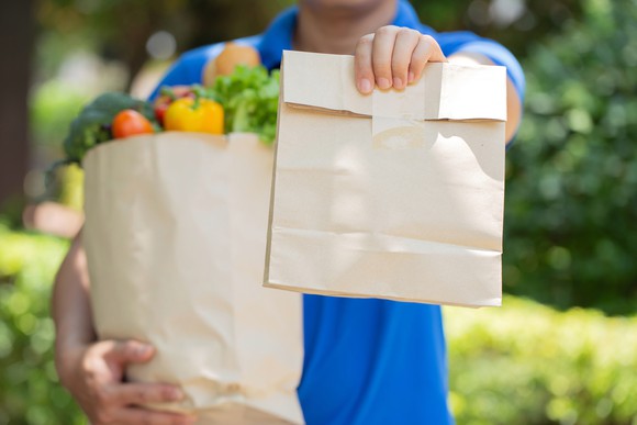 A delivery man seen from the neck down carrying groceries and holding out a sealed paper bag.