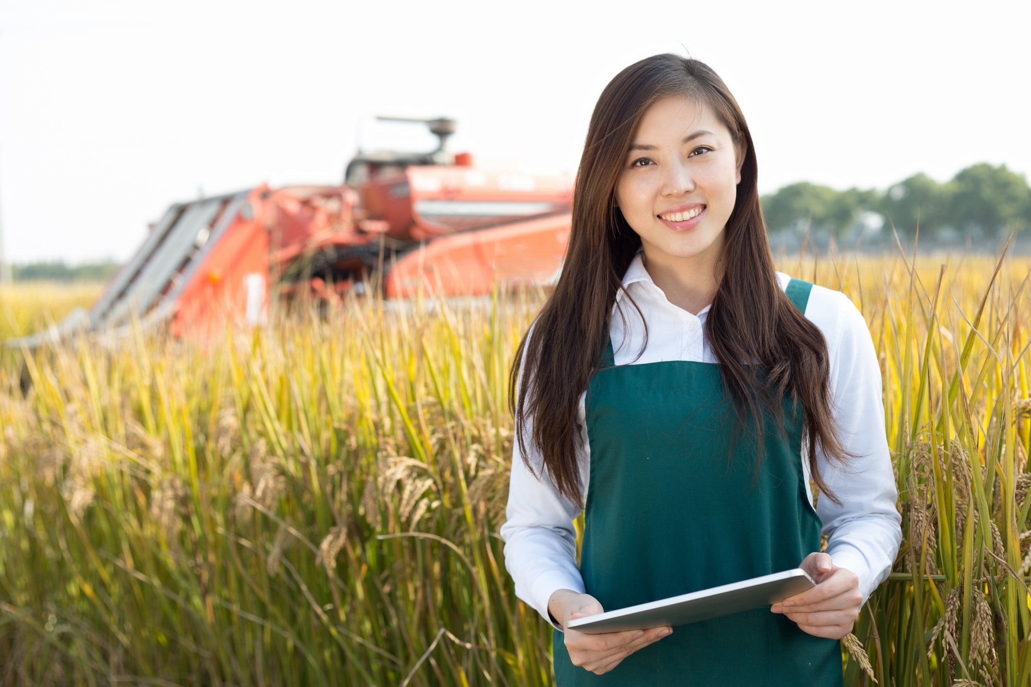 A young woman smiles holding a tablet in a field in front of farm equipment. 