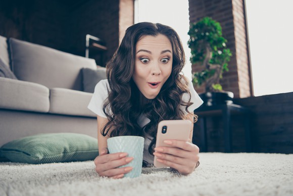 A young woman lies prone on the floor and is shocked by something on her smartphone.