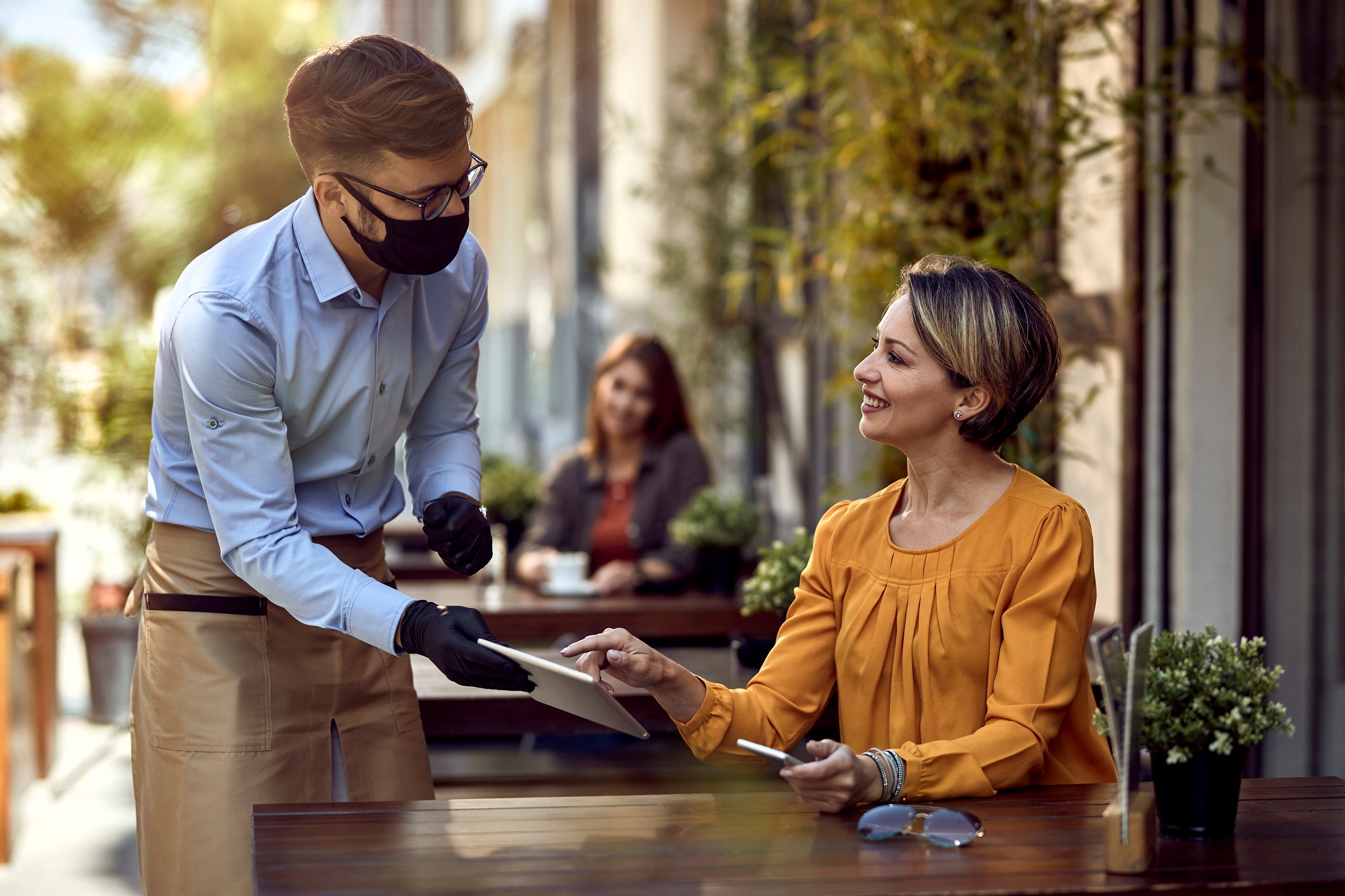 A waiter with a face mask and gloves and a woman ordering on a tablet in a restaurant