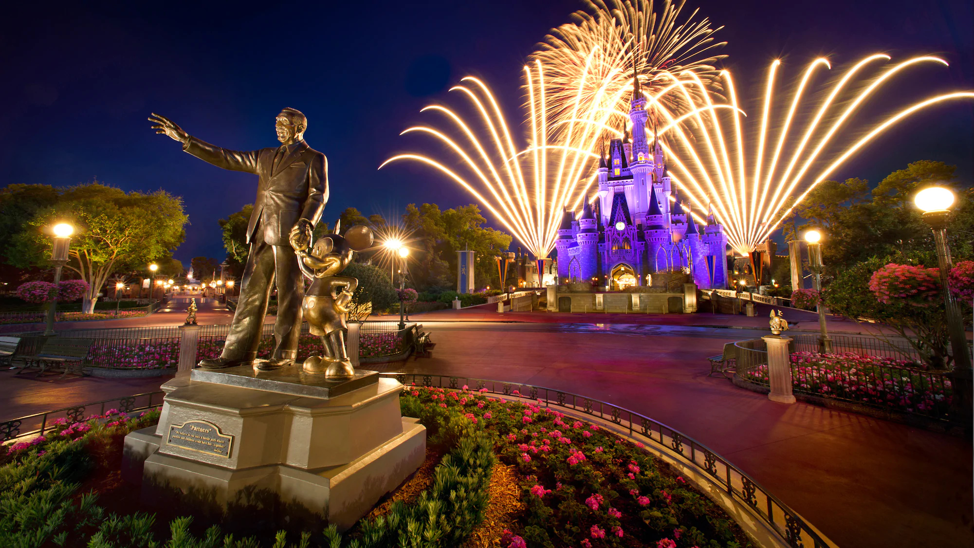 Fireworks at night over the Cinderella Castle and a bronze statue of Mickey and Walt Disney.