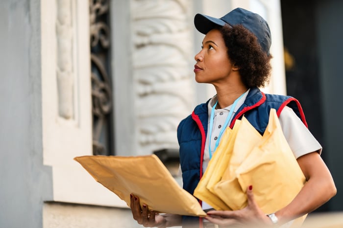 Person wearing blue and red vest carrying envelopes in front of a house.