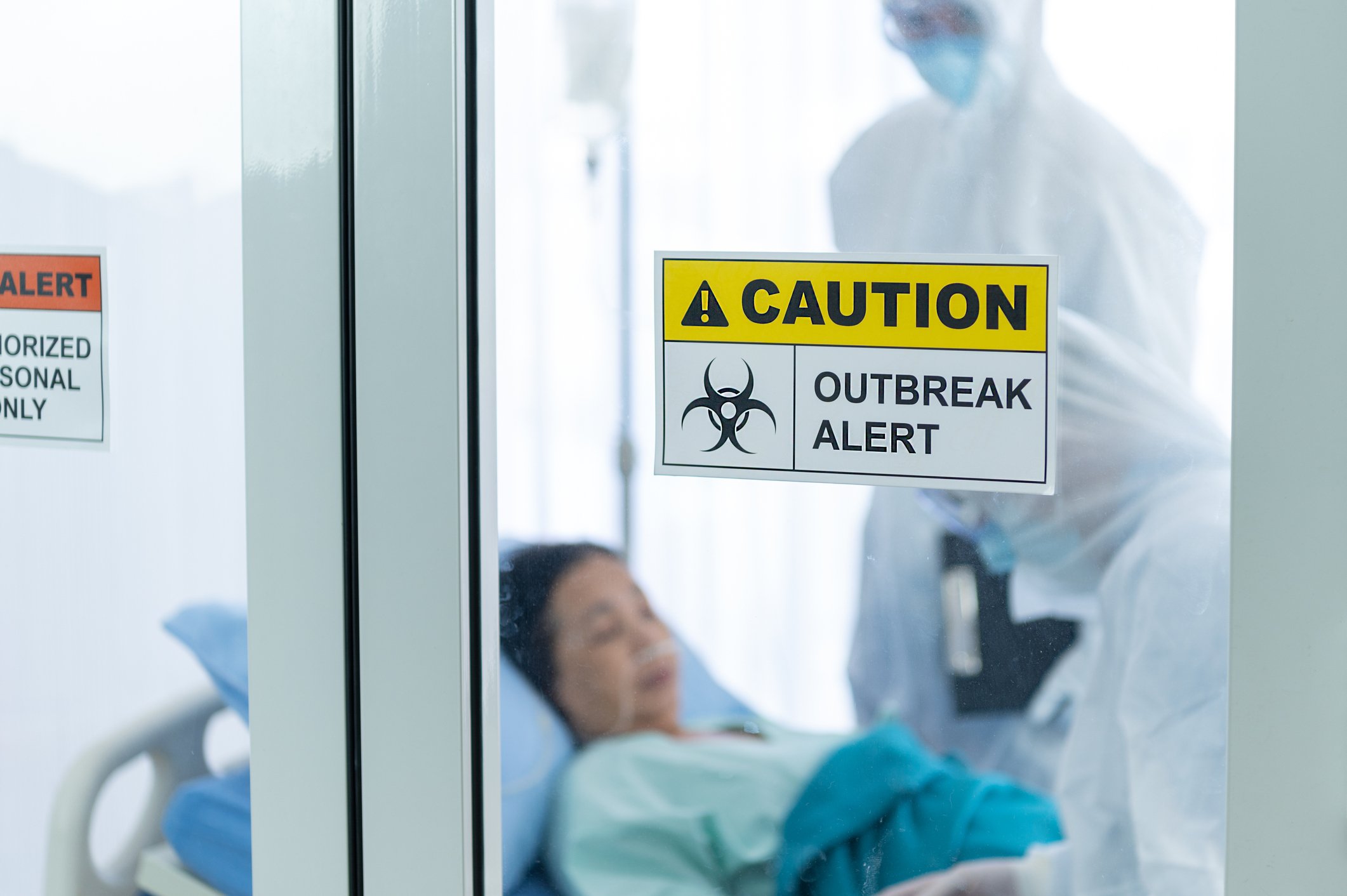 Doctors in personal protective equipment and a patient behind a glass door with a sign that reads caution: outbreak alert. 