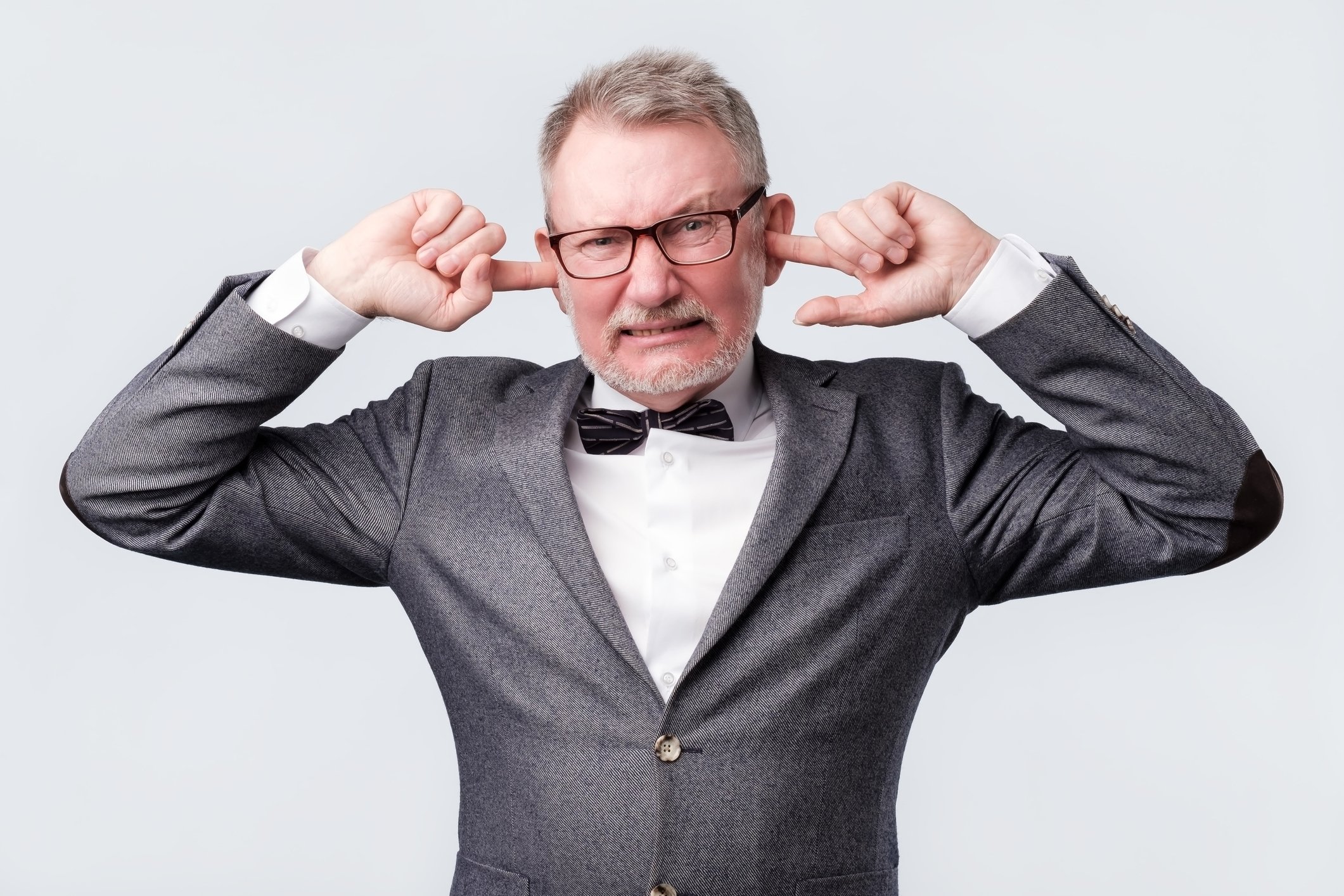 An old man in a grey suit plugs his ears with his index fingers.