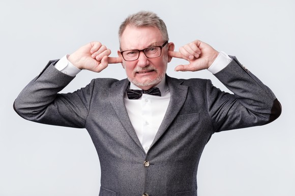 An old man in a grey suit plugs his ears with his index fingers.