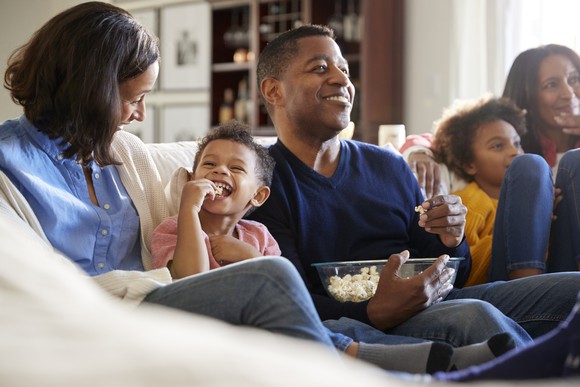 Family sitting on a sofa while watching a movie and eating popcorn.