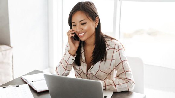 A young woman at home on a phone working on a laptop in pajamas.