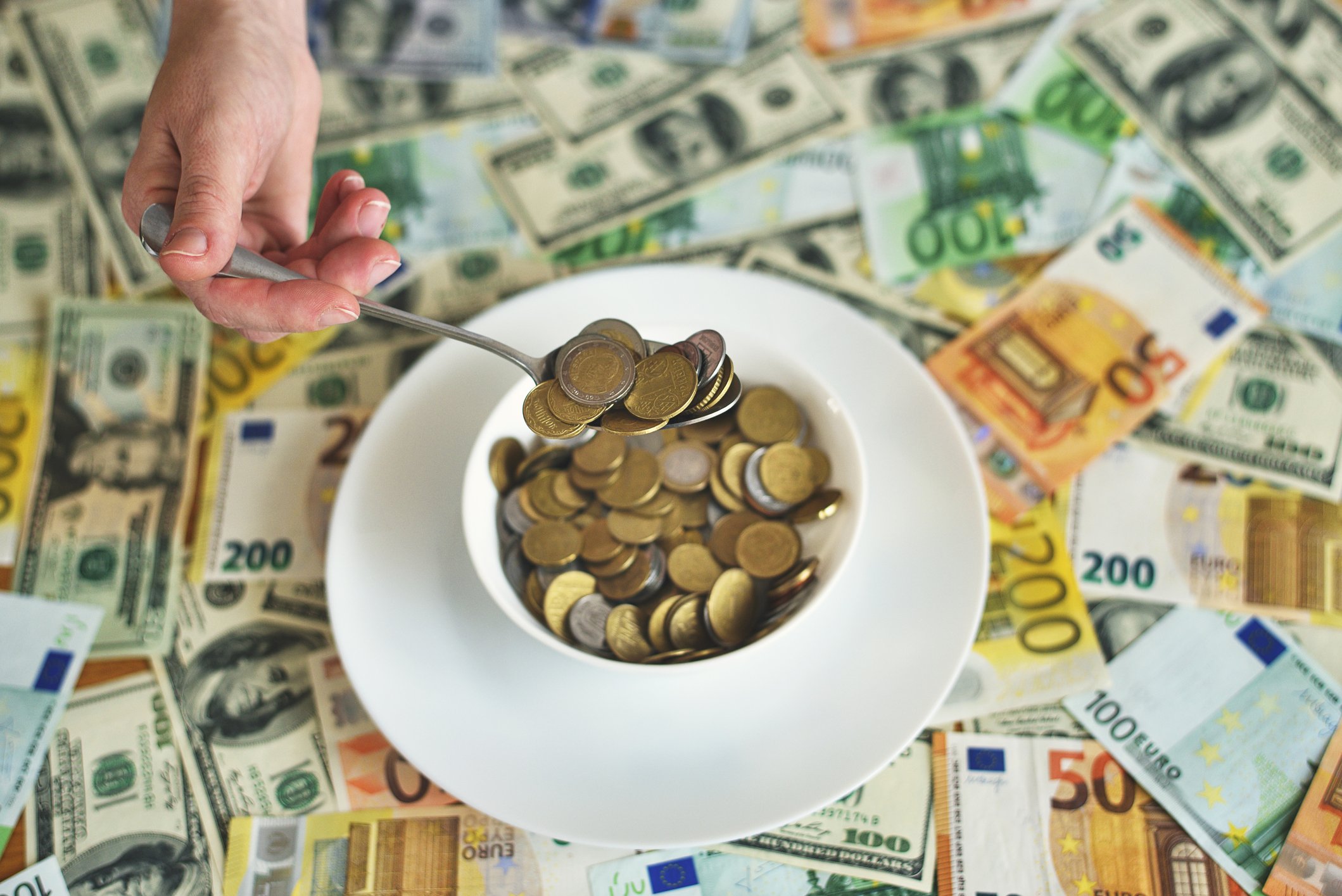 Coins in bowl being scooped out by spoon with various currency underneath